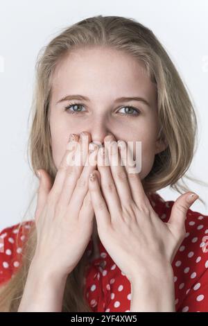 Blonde Frau bedeckt ihren Mund mit ihren Händen und schaut in die Kamera. Porträt eines 21-jährigen, authentischen weiblichen Models, gekleidet in sommerroter Polka Dot dre Stockfoto