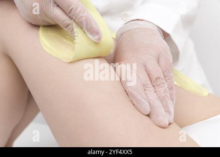 Nahaufnahme von Kosmetikhänden, die Frauenbeine mit grünem heißem Wachs im Schönheitssalon enthaeben. Der Wachsvorgang der Haarentfernung aus der Wurzel. Frauen inti Stockfoto