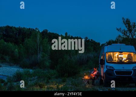 Ein Mann sitzt auf einem Campingstuhl vor einem Tisch an einem Sommerabend zur blauen Stunde vor seinem Camper. Deutschland mit einem Campervan unterwegs *** Ein Mann sitzt auf einem Campingstuhl vor einem Tisch an einem blauen Stunden Sommerabend vor seinem Campervan Deutschland auf der Straße in einem Campervan Stockfoto