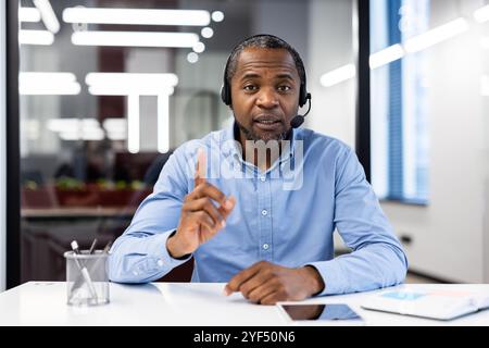 Reifer afroamerikanischer Geschäftsmann, der während einer Videokonferenz im modernen Büro ein Headset trägt. Gespräche mit ausdrucksstarken Gesten führen, die sich auf Kommunikation und Teamarbeit konzentrieren. Stockfoto