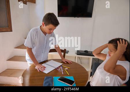 Eine Mutter hilft ihrem Sohn bei seinen Hausaufgaben an einem runden Küchentisch. Der Junge ist konzentriert, während er Notizen in ein Notizbuch schreibt, umgeben von stationärem und Stockfoto