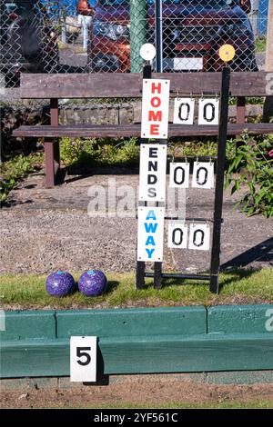 Eine Anzeigetafel für Bowls, die auf einem Bowlinggrün steht, vor einer hölzernen Zuschauerbank. Ein paar lila Schüsseln liegen auf dem Gras Stockfoto