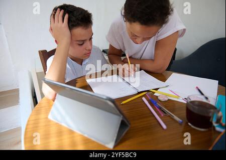 Eine Mutter hilft ihrem Sohn mit Hausaufgaben an einem Tisch, umgeben von Studienmaterialien und einem Tablet. Sie konzentrieren sich auf die gemeinsame Lösung von Problemen. Stockfoto