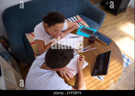 Eine Mutter hilft ihrem Sohn bei seinen Hausaufgaben in einem gemütlichen Zuhause. Sie sind umgeben von Notebooks, einem Tablet und einer Tasse Kaffee, die den Fokus betonen Stockfoto