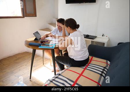 Zwei Frauen konzentrieren sich intensiv auf die Arbeit und arbeiten zu Hause an einem Projekt zusammen. Sie sitzen an einem Tisch mit einem Laptop und Dokumenten, vermitteln Teamarbeit und Konzentration Stockfoto