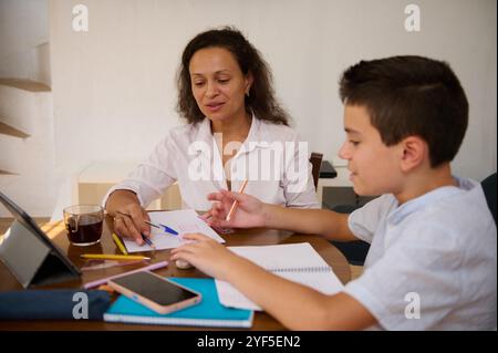 Eine Mutter hilft ihrem Sohn bei den Hausaufgaben, während sie an einem Tisch sitzt. Sie verwenden ein Tablet, umgeben von Notizbüchern und Bleistiften, was eine Unterstützung fördert Stockfoto