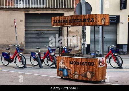 Viladecans. Spanien - 03. November 2024: Ein hölzerner Straßenwagen, der geröstete Kastanien und Süßkartoffeln verkauft, befindet sich auf einem Bürgersteig mit Fahrrad Stockfoto