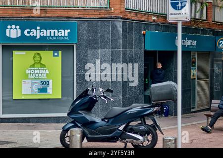 Viladecans. Spanien - 3. November 2024: Ein moderner schwarzer Roller steht vor einer ländlichen Bank namens Cajamar mit einem Werbeplakat im Fenster und Stockfoto