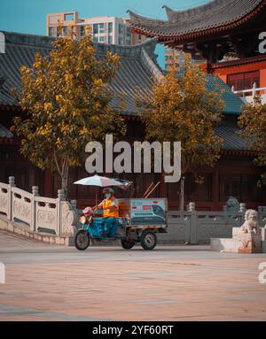 Ein Sanitär-Mitarbeiter auf einem dreirädrigen Nutzfahrzeug, das mit einem Sonnenschirm ausgestattet ist, fährt vorbei an traditioneller chinesischer Architektur Stockfoto