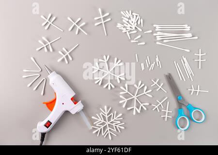Handgefertigte Baumwoll-Wattestäbchen mit Schneeflocken-Dekorationen auf grauer Oberfläche mit Heißklebepistole und Schere daneben. Flache Ladefläche mit Ideen für das Basteln im Winter und Stockfoto