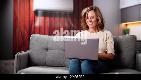 Junge Glückliche Frau Unter Klimaanlage Auf Der Couch Mit Laptop Zu Hause Stockfoto