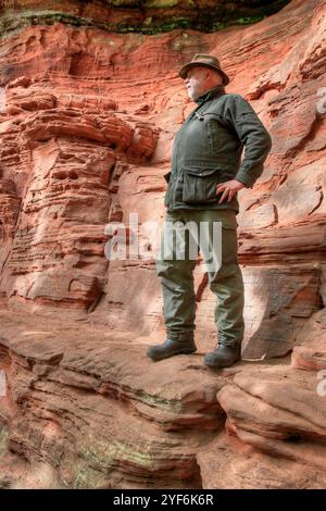 Ein älterer Wanderer steht am Rande eines schmalen felsigen Pfades mit einer steilen Sandsteinklippe hinter ihm. Stockfoto