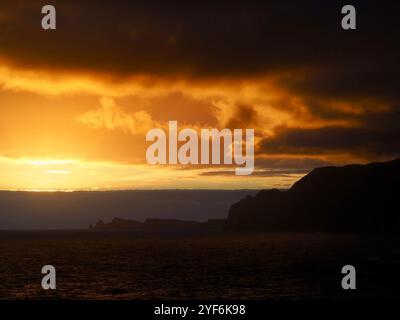 Der Himmel brennt mit feurigen Tönen, wenn die Sonne untergeht, und strahlt ein orangenes Leuchten über einer zerklüfteten Küste aus, das einen scharfen Kontrast zum dunklen Ozean unter Madeira bildet Stockfoto