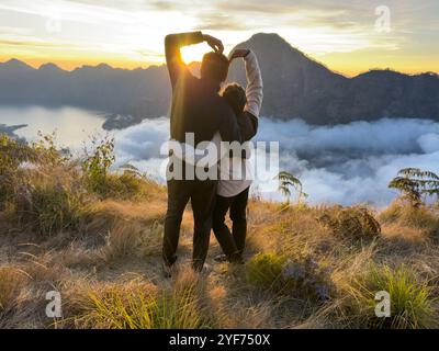 Rückansicht eines Paares, das ein Herzzeichen macht, während er den Sonnenuntergang auf einem Kletterausflug beobachtet, Mount Rinjani, Lombok, West Nusa Tenggara, Indonesien Stockfoto