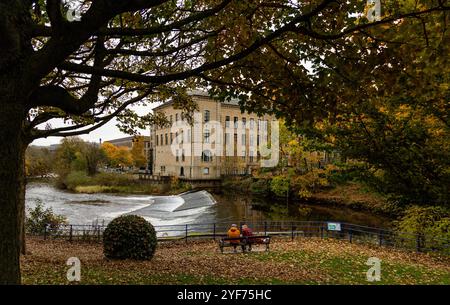 Blick auf Salts Mill, Saltaire vom Roberts Park in Baildon im Herbst. Der Fluss Aire fließt über ein Wehr zwischen Mühle und Park. Stockfoto