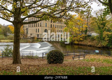 Blick auf Salts Mill, Saltaire vom Roberts Park in Baildon im Herbst. Der Fluss Aire fließt über ein Wehr zwischen Mühle und Park. Stockfoto