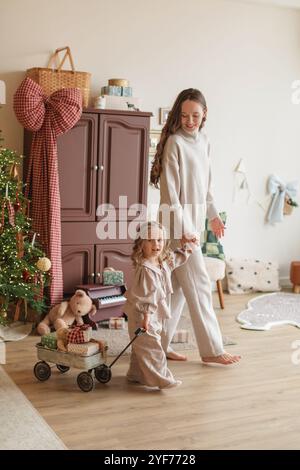 Lächelnde Mutter und Tochter gehen Hand in Hand und ziehen einen Wagen mit Weihnachtsgeschenken in ein Schlafzimmer Stockfoto