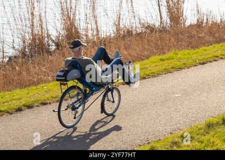 Ein älterer Mann fährt mit Schilf auf einem Liegefahrrad am Wasser entlang. Stockfoto