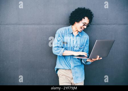 Ein junger Mann mit lockigen Haaren, der ein blaues Jeanshemd trägt, steht vor einer dunkelgrauen Wand, hält einen Laptop in der einen Hand und tippt mit der anderen, lächelnd Stockfoto