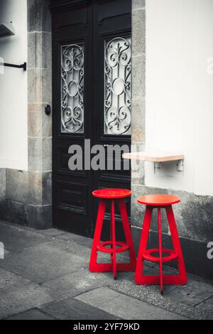 Vertikale Aufnahme von zwei roten Hockern vor einem Straßencafé. Die Hocker sind an eine weiße Wand mit einer schwarzen, verzierten Tür mit dekorativem Eisen gestellt. Stockfoto