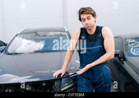 Verwirrter Mechaniker in blauem Overall, der neben dem beschädigten Auto steht und die Motorhaube mit traurigem und besorgtem Gesichtsausdruck berührt. Stockfoto