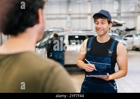 Positiver Mechaniker in Uniform erklärt Checkliste Fahrzeugwartung und -Reparatur vor nicht erkennbarem man-Kunden in der Fahrzeugwerkstatt. Stockfoto