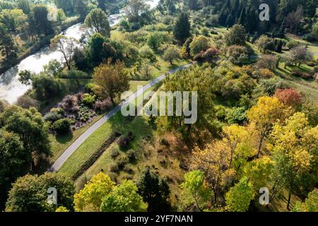 Ein Blick aus der Vogelperspektive fängt einen botanischen Garten mit lebhaften Herbstbäumen, einem gewundenen Pfad und einem ruhigen Fluss ein, umgeben von dichtem Wald. Stockfoto