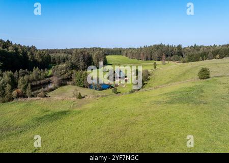 Eine ruhige ländliche Szene bietet ein kleines Haus mit einem grünen Dach, umgeben von sanften Hügeln, einem ruhigen Teich und üppigen Bäumen unter einem klaren blauen Himmel. Stockfoto