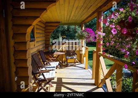 Eine Blockhüttenveranda verfügt über rustikale Holzstühle, einen runden Tisch und farbenfrohe hängende Blumenkörbe vor einer üppigen grünen Gartenkulisse. Stockfoto