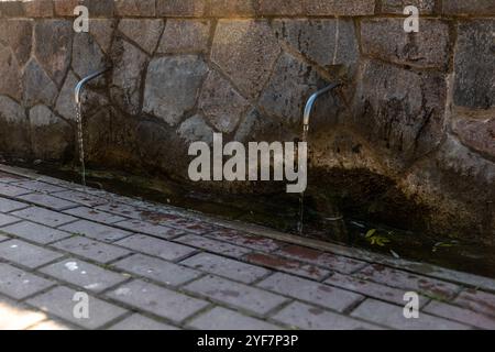 Eine Steinmauer verfügt über zwei Metallausläufe, in denen Wasser in einen kleinen Bach fließt. Sonnenlicht hebt die Textur der Steine und Ziegelpflaster darunter hervor. Stockfoto