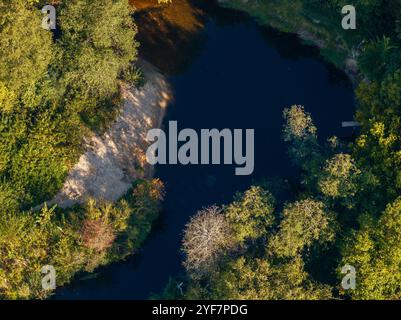 Aus der Vogelperspektive fängt ein kleiner, sich windender Fluss mit dunklem Wasser ein, umgeben von lebendigem Grün und einer sandigen Bank auf der linken Seite. Stockfoto