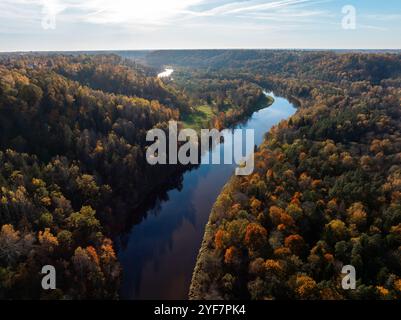 Der Fluss Gauja schlängelt sich durch einen dichten Wald in Sigulda, Lettland, umgeben von lebhaftem Herbstlaub. Hügel und ein klarer blauer Himmel runden die Szene ab. Stockfoto