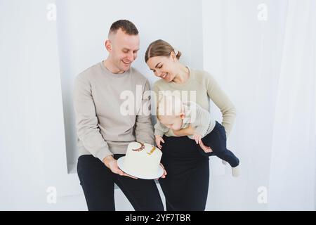 Lächelnde Mutter und Vater halten ein Kind und einen Urlaubskuchen. Eltern, Mom und Dad und lächelndes Baby in den Armen isoliert über weißem Hintergrund. Fröhlich, jung Stockfoto