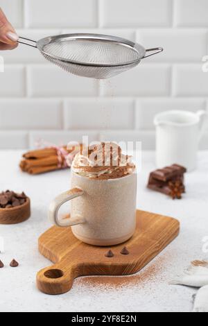 Kakaopulver auf Schlagsahne in der Tasse auf dem Schneidebrett gießen Stockfoto