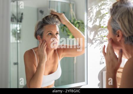 Europäische schöne Frau mittleren Alters untersucht sorgfältig ihre grauen Haare und Falten im Spiegel. Stockfoto