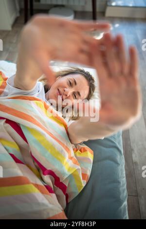 Porträt Frau mittleren Alters durch Palme lächelnd unter Decke im Bett wach mit guter Laune. Stockfoto
