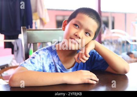 Ein kleiner Junge sitzt nachdenklich an einem Tisch und legt sein Kinn auf seine Hand. Sein ausdrucksstarkes Gesicht spiegelt Neugier und Nachsicht in einem zwanglosen Innenbereich wider Stockfoto