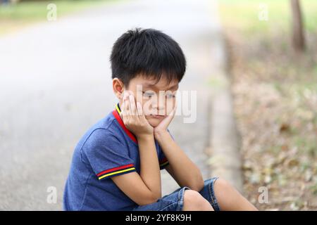 Ein kleiner Junge sitzt allein auf einem Weg, legt sein Kinn auf seine Hände, verloren in Gedanken. Dieser emotionale Moment fängt das Wesen der kindlichen Kontempla ein Stockfoto