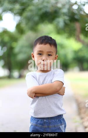 Ein selbstbewusster kleiner Junge posiert mit überkreuzten Armen in einem sonnigen Park, umgeben von üppigem Grün. Sein Ausdruck fängt Unschuld und Verspieltheit auf einem bea ein Stockfoto