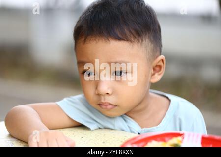 Ein nachdenklicher kleiner Junge sitzt an einem Tisch im Freien, verloren in Reflexion, während er ein Essen genießt. Sein ernster Ausdruck fängt einen Moment der Unschuld und c ein Stockfoto