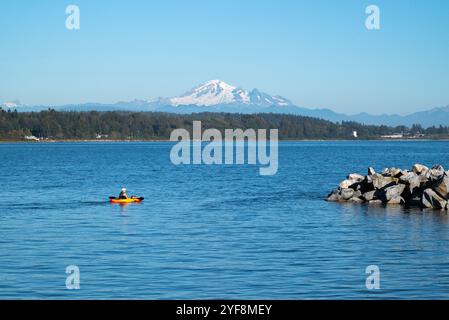 Ein Mann in einem gelben Kajak paddelt entlang des Ufers von White Rock mit dem Berg im Hintergrund. Stockfoto