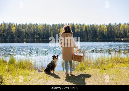 Eine junge Frau mit dem Hund Australian Shepherd hält einen Picknickkorb in der Hand vor dem Hintergrund des Sees Stockfoto