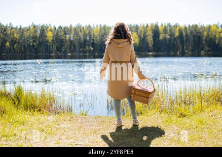 Die junge Frau hält einen Picknickkorb in der Hand vor dem Hintergrund des Sees Stockfoto