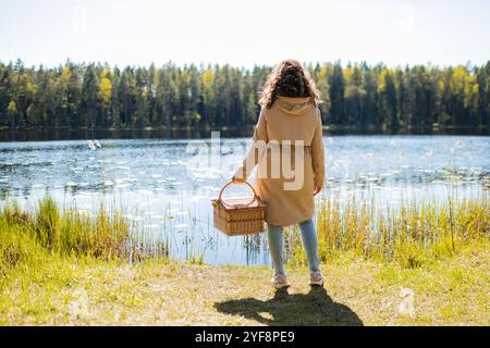 Die junge Frau hält einen Picknickkorb in der Hand vor dem Hintergrund des Sees Stockfoto