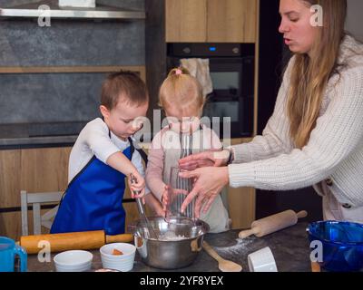 Mom und ihre beiden Kinder machen Keksenteig in einer modernen Küche, indem sie sorgfältig Zutaten in einer Schüssel mischen. Lächelnd, während sie rühren, die Kinder Stockfoto