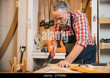 Ein Zimmermann in rotem kariertem Hemd und blauem Overall sägt ein Stück Holz mit einer Handsäge auf einem Stockfoto