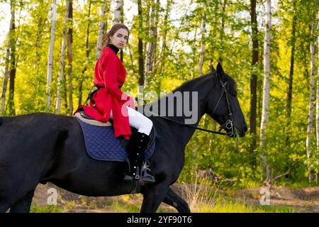 Die Frau reitet auf einem schwarzen Pferd im Wald. Die Frau trägt einen roten Mantel und eine weiße Hose. Das Pferd ist schwarz und hat einen schwarzen Sattel. Der Wald ist l Stockfoto