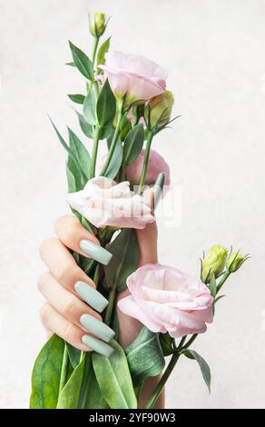 Eine schlanke Hand zeigt lange, pastellfarbene Nägel, während sie sanft einen Blumenstrauß aus zarten rosa Blüten ergreift, was eine ruhige und elegante Ästhetik hervorhebt. Stockfoto