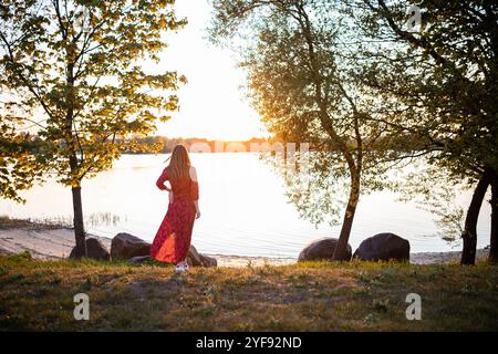 Frau in rotem Kleid genießt einen friedlichen Sonnenuntergang am Seeufer, das goldene Sonnenlicht strahlt ein warmes Licht auf das ruhige Wasser Stockfoto