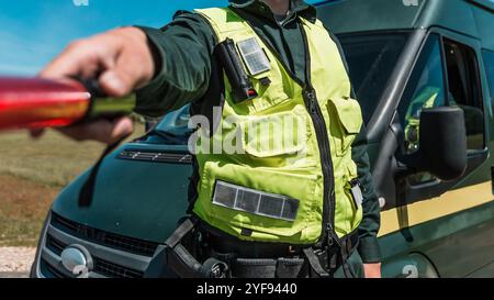 Verkehrsoffizier in einer Warnweste mit einem Stab zur Straßenkontrolle in der Nähe eines Patrouillenfahrzeugs, mit Sicherheitsausrüstung Stockfoto